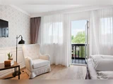 Et lyst, hyggeligt hotelværelse med en stribet lænestol, natbord, stor seng, væghængt tv og åbne altandøre, der lukker naturligt lys ind og giver udsigt til de grønne områder udenfor.
