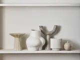 Fem moderne keramikvaser og -kar i forskellige abstrakte former og neutrale toner står på en hvid hylde mod en lys væg.