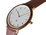 Et minimalistisk armbåndsur med hvid urskive, timemarkeringer og visere i guld, en brun læderrem og en urkasse i guld. Tiden viser ca. 10:10.