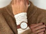 En person iført en solbrun sweater og et hvidt hæklet ærme viser et guldkantet armbåndsur med en brun læderrem på sit håndled.