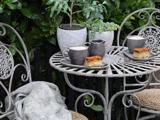 Et havebord i smedejern med to kopper kaffe, to tallerkener med kage, en mælkekande og potteplanter, omgivet af grønne blade.