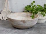 En rustik, lys træskål står på en grå flade ved siden af en hvid, struktureret potte med grønne urter og to træredskaber i baggrunden.