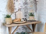 Et rustikt træbord med brød, ost, potteplanter, en sort-hvid ko-figur og dekorative skærebrætter mod en hvid murstensvæg.