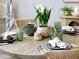 Et rundt træbord dækket til fire med grønne glas, neutrale tallerkener, stofservietter og en vase med hvide blomster som midterstykke. Et stearinlys og en potteplante står i baggrunden.