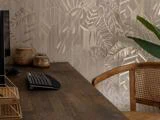 Et træskrivebord med tastatur, skærm og flettede kurve står ved siden af en rottingstol. Baggrunden er et beige, bladmønstret tapet og en stor grøn plante.