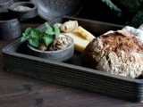 En rustik bakke med et brød, en lille skål med friske krydderurter og rødder og skiver af ost på et træbord.
