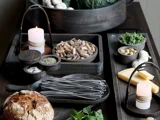 På et rustikt bord står pasta, krydderurter, brød, ost, hvidløg, krydderier, bladgrønt og et tændt stearinlys i hyggelige, jordnære omgivelser.