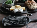 Sort pasta, to hvidløg, osteskiver og hakket grønt ligger på rustikke træfade ved siden af et rundt brød på et træbord.