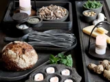 En rustik bordopstilling med sorte bakker med pasta, brød, ost, grønt, nødder, hvidløg, krydderier og tændte stearinlys skaber en hyggelig og indbydende atmosfære.