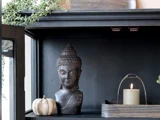 I et sort skab står en statue af et buddha-hoved, et lille hvidt græskar og en bakke med et tændt beige lys og grønne planter.