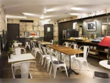 Moderne café med hvide stole, træborde, vægkunst, potteplanter og en morgenmadsbuffet med kager, drikkevarer og frugt langs bagvæggen. Varm belysning skaber en hyggelig atmosfære.