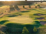 Luftfoto af en solbeskinnet golfbane med grønne fairways, sandbunkers og en enkelt person på en plæneklipper i midten blandt træer og gyldent græs.
