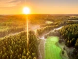 Luftfoto af en frodig grøn golfbane omgivet af tæt skov ved solnedgang, hvor sollyset strømmer ind over landskabet.