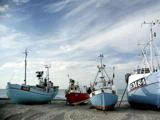 Flere fiskerbåde med antenner er strandet på sandet under en delvist overskyet himmel med havet synligt i baggrunden.