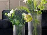 To klare, riflede glasvaser indeholder grønne og hvide blomster med grønne blade på en mørk, sløret baggrund.