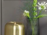En struktureret, mørk vase med hvide og grønne blomster står ved siden af en rund, guldfarvet dekoration på en grå væg.