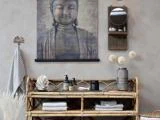 En bambushylde med håndklæder, toiletartikler og dekorationer står under et stort Buddha-kunstværk; et rundt spejl og en væghylde hænger i nærheden og skaber en fredfyldt, spa-agtig atmosfære.