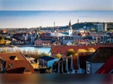 Et levende bybillede med bygninger med røde tage, et fremtrædende kirketårn, træer og vand i baggrunden under en klar blå himmel.