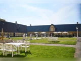 En gårdhave med hvide udendørsborde og -stole, grønt græs, statuer, et springvand og en stor gul bygning i baggrunden under en klar himmel.