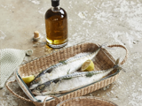 To hele rå fisk med citronbåde og krydderurter i en flettet kurv ved siden af en flaske olivenolie på en rustik overflade.