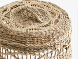 Nærbillede af en flettet skammel eller kurv i rattan eller søgræs med et spiralmønster på den flade top og et luftigt, åbent vævet design på siderne.