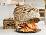 En hånd løfter en flettet kurvs madlåg fra en tallerken med tre croissanter på et bord. En anden flettet kurv ses i baggrunden.