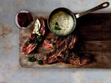 En træplade med krydrede kødstrimler, en lille gryde med cremet sauce, en skål med skiveskårne rødbeder og pyntet med friske krydderurter.