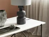 Et moderne hvidt bord med en mørk skulpturel lampe, en keramikvase og en bakke med garn og saks. Lyse gardiner hænger i baggrunden.
