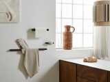 Et minimalt badeværelse har en håndklædestang med et håndklæde og en avis, en væghængt toiletpapirholder, et vindue med en keramisk vase og en bordplade med brød.