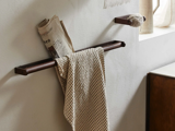 Et mønstret håndklæde hænger på en brun håndklædestang med en sammenrullet avis ovenpå; en toiletpapirrulle ligger på en holder, der er monteret på væggen i nærheden.