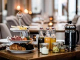 Morgenmad på et træbord med kager, oste, pålæg, glas med juice, frisk frugt, marmelade, mælk og en fransk kaffemaskine i et hyggeligt spisemiljø.