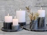 Fire hvide lys i forskellige højder brænder i runde, mørke marmorstager ved siden af en matchende vase med tørret græs, alt sammen placeret på stenbakker mod en grå struktureret baggrund.