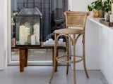 En flettet barstol står ved en hvid disk; i nærheden holder en træbænk en glaslanterne med tændte stearinlys og grønne planter i et hyggeligt, moderne rum.