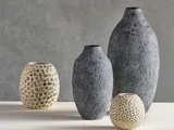 Fire teksturerede vaser i forskellige størrelser, to mørkegrå og to cremefarvede med prikkede mønstre, er udstillet på en lys overflade mod en grå baggrund.