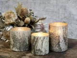 Tre rustikke, riflede glaslysestager med tændte lys indeni er placeret på en træoverflade ved siden af et tørret blomsterarrangement mod en struktureret grå væg.