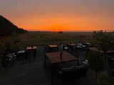 En udendørs terrasse med borde og stole med udsigt over en mark ved solnedgang.