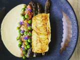 En gourmetret med stegt fisk, asparges, grønne ærter, spiselige blomster og sauce, elegant anrettet på en mørkeblå keramiktallerken.