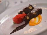 En anrettet dessert med to stykker chokolade- og nøddebrownie, friske jordbær i skiver, appelsinsauce og et drys chokolade på en hvid tallerken.