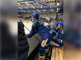Ishockeyspillere i blå trøjer sidder og står i holdets bænkområde under en kamp i en indendørs arena. Der ses tilskuere i baggrunden.