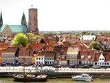 En udsigt over vandet i en dansk by med huse med røde tage, kirketårne, både, der ligger til kaj, og mennesker, der går langs vandet.