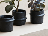 Tre sorte, teksturerede urtepotter, to med grønne planter og en tom, står på en lys overflade mod en neutral væg.