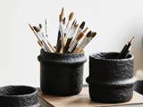 Tre sorte beholdere med tekstur holder pensler, blyanter og andre kunstartikler på en træoverflade mod en almindelig baggrund.
