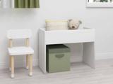 Et lille hvidt skrivebord med en grøn opbevaringsboks under, en pude og en bamse ovenpå, ved siden af en hvid træstol i et lyst, minimalistisk rum.