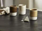 Tre keramiske kopper og en pyramideformet tepose står på en mørk overflade med en beige serviet i baggrunden.