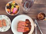 Mediumstegt bøf i skiver med sauce på en tallerken, en frisk salat, brødskiver, et glas rødvin og en lille skål med sauce på et træbord.
