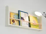 En hvid væghængt bogreol rummer tre børnebøger og oplyses af en hvid lampe, der er monteret på væggen til højre.