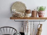 En træhylde med flettede kurve, lerkrukker, træskåle, en potteplante og køkkenredskaber hængende under; en stol er delvist synlig til venstre.