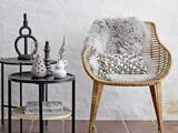 En kurvestol med en fluffy grå plaid og en struktureret pude står ved siden af to sorte sideborde med vaser, dekorationer og en bog på en hvid struktureret væg og et flisegulv.