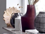 Nærbillede af forskellige dekorative vaser på en marmorhylde, bl.a. en stor vinrød vase, en hvid vase, en sort vase og en sort lanterne med struktur.