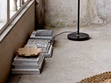 Stablede bøger med en dekorativ skål på toppen står på et betongulv ved siden af en sort gulvlampe og en struktureret væg.